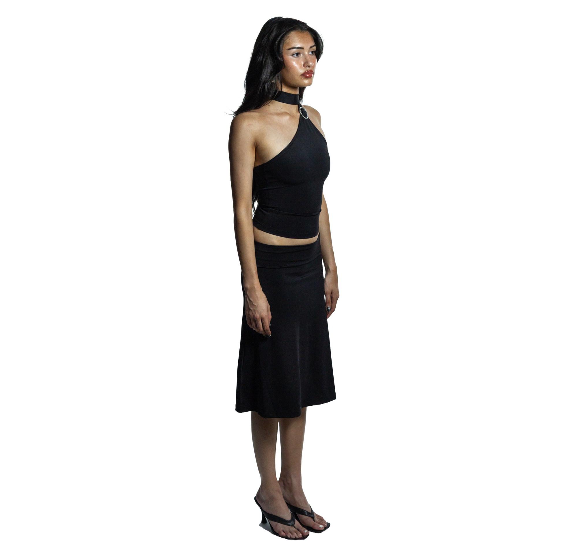 Full body side view of jet black self-tieing asymetrical top with circle brooch and midi knee-length skirt